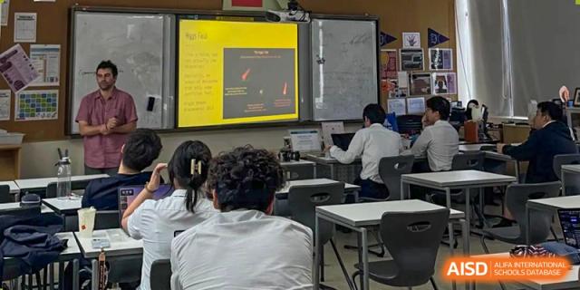 Students looking at a digital presentation displayed on a screen at the front of the classroom during a lesson, with the teacher standing to the side, at Shanghai United International School, Wanyuan Campus.
