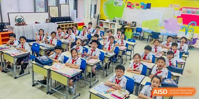 A wide shot of a brightly lit elementary or middle school classroom filled with young students sitting at desks at Shanghai United International School, Wanyuan Campus.