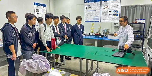 Three male students in suits and ties standing in a lab or workshop near a green table, possibly for a science or engineering competition at Shanghai United International School, Wanyuan Campus.