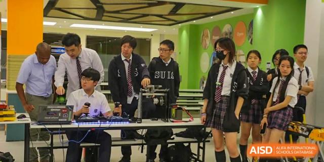 Students surrounding a device or robot on a table, possibly for a science or technology club demonstration, with one student wearing a BULLS sweater at Shanghai United International School, Wanyuan Campus.