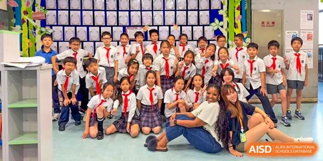 A large group of young students in uniform and red scarves kneeling and sitting for a group photo in front of a wall of student-made artwork or awards at Shanghai United International School, Wanyuan Campus.