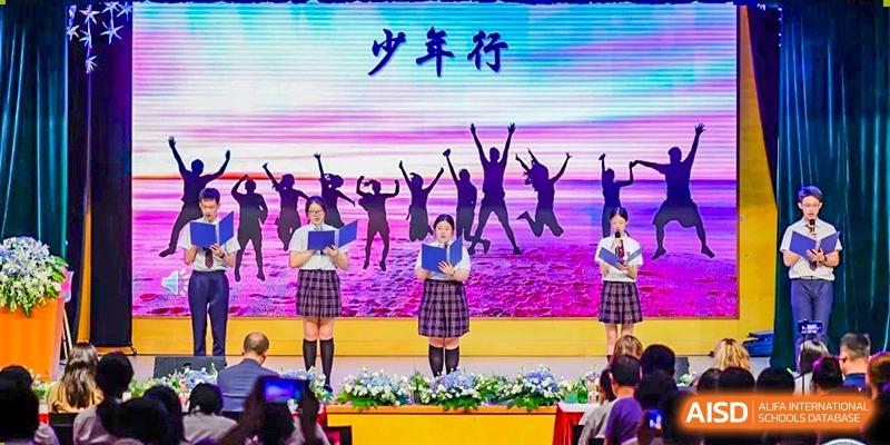 Students in uniform performing on stage with a screen behind them showing jumping silhouettes and the Chinese title "少年行" (Youth Journey) at Shanghai United International School, Wanyuan Campus.