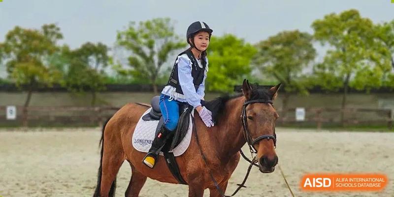 A young student wearing a riding helmet and vest, sitting on a brown horse outdoors during an equestrian club activity at Shanghai United International School, Wanyuan Campus.