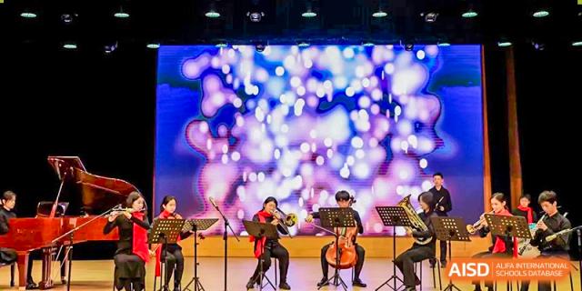 Musicians playing string instruments, a cello and a violin, on a stage with a dazzling, out-of-focus background of bright blue and white lights at Shanghai United International School, Wanyuan Campus.