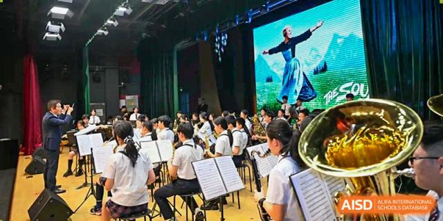 Students in an orchestra or band playing their instruments in the foreground, with a performer on a brightly lit stage in the background, during an assembly or concert at Shanghai United International School, Wanyuan Campus.
