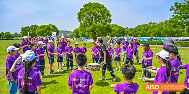 A group of students in purple shirts playing drums outdoors in a field, led by a teacher in a black shirt, at Shanghai United International School, Wanyuan Campus.