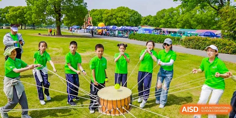 Students in matching green shirts participating in a team-building or traditional drumming activity outdoors at Shanghai United International School, Wanyuan Campus.