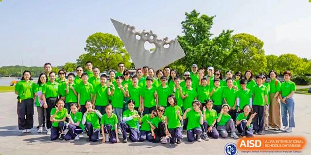 A large group of students in bright green shirts posing outdoors on the grass in front of an abstract gray sculpture at Shanghai United International School, Wanyuan Campus.