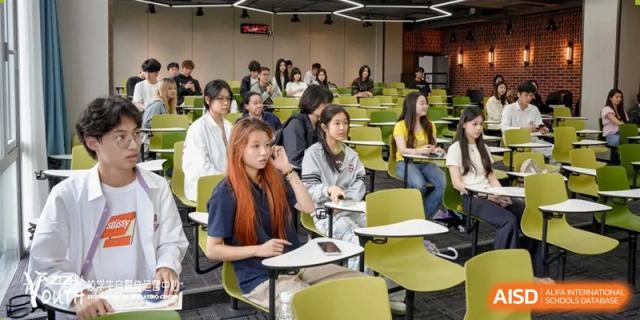 A classroom scene at SHENGHUA ZIZHU ACADEMY with students seated at green desks, participating in a lecture or discussion.