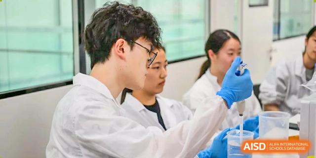 A close-up of a male student from SHENGHUA ZIZHU ACADEMY wearing glasses and a lab coat, looking intently at a demonstration or experiment.