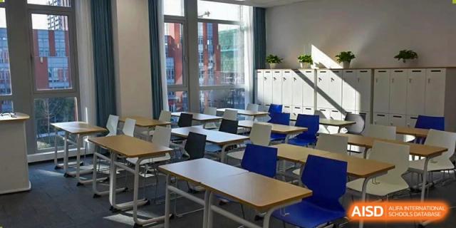A classroom at SHENGHUA ZIZHU ACADEMY with rows of wooden desks, blue chairs, and metal lockers visible along the back wall.