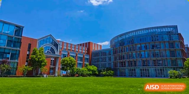 The exterior of the modern academic buildings of SHENGHUA ZIZHU ACADEMY, featuring red-brick and glass facades, surrounded by a bright green lawn under a blue sky.