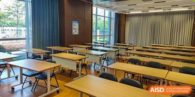 A bright, modern classroom at SHENGHUA ZIZHU ACADEMY with rows of individual desks and chairs next to large windows overlooking a green area.