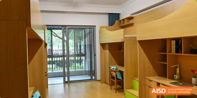 An interior view of a dormitory room at SHENGHUA ZIZHU ACADEMY, featuring a wooden loft bed, desk, and a glass door leading to a balcony.