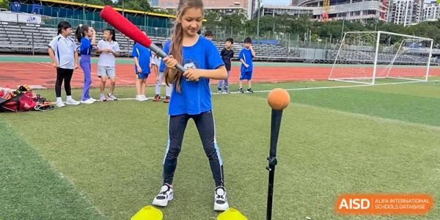 A girl in a blue shirt standing ready with a bat near a batting tee on a sports field at Shenzhen American International School.