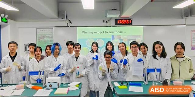 A group of smiling students in lab coats posing for a photo in a science class with a presentation screen visible at Shenzhen College of International Education.