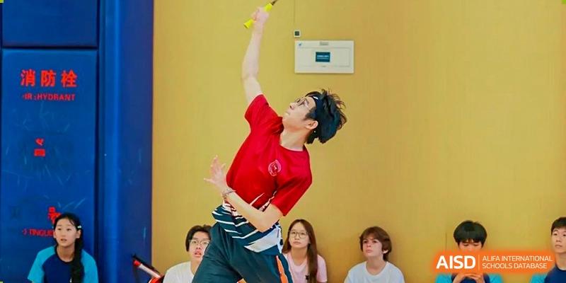 A male student in a red shirt hitting a shuttlecock during a badminton match at Shenzhen College of International Education.