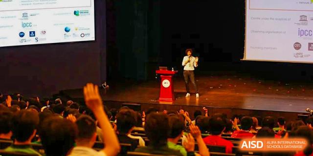 A student speaking into a microphone on a stage during a school event or performance to an engaged audience at Shenzhen College of International Education.