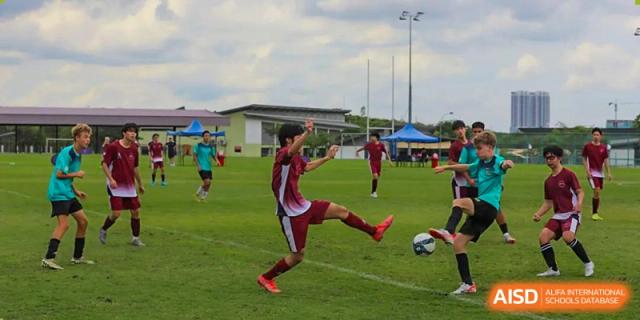 A student in a red and white uniform kicking a soccer ball during a game on a green field at Shenzhen College of International Education.