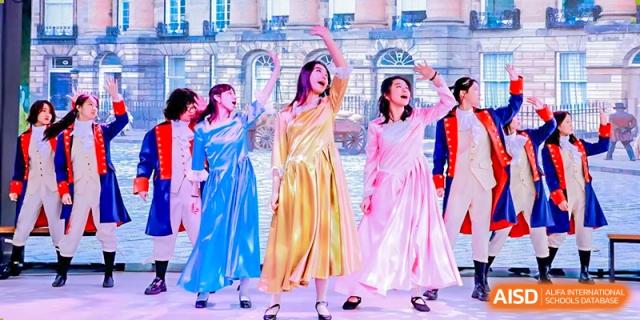 Students wearing colorful blue, yellow, and pink gowns performing a dramatic dance or theatrical piece on a stage at Shenzhen International Foundation College (SIFC).