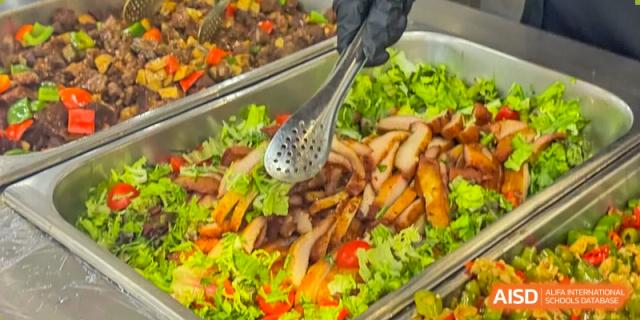 A close-up of a serving spoon over a healthy buffet dish with grilled meat and fresh salad at Shenzhen International Foundation College (SIFC).