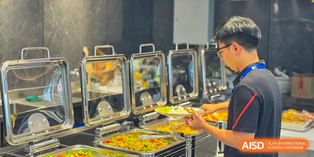 A student or staff member serving themselves from a buffet line with multiple hot food trays in the cafeteria at Shenzhen International Foundation College (SIFC).