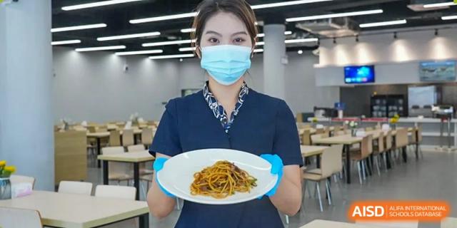 A staff member wearing a face mask and gloves holding a plate of pasta in the school cafeteria at Shenzhen International Foundation College (SIFC).
