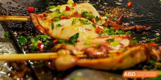 A close-up of a grilled fish fillet topped with colorful peppers and herbs being prepared for a school meal at Shenzhen International Foundation College (SIFC).