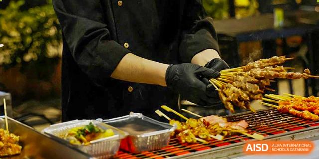 A chef wearing black gloves grilling skewers of meat and vegetables for students at Shenzhen International Foundation College (SIFC).