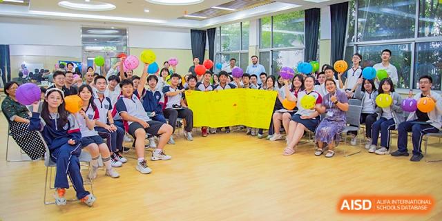 A large group of Suzhou Innovation Academy students seated in a brightly lit room holding balloons and a yellow banner for a classroom or club event.