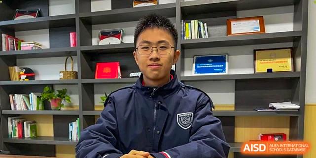 A male student from Suzhou Innovation Academy wearing a navy blue school jacket and glasses posing for a portrait in front of a bookshelf.