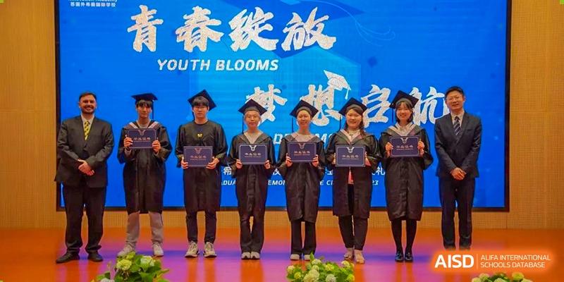 Four graduating students from Suzhou Innovation Academy in caps and gowns holding their diplomas on stage at a ceremony with a "YOUTH BLOOMS" banner.
