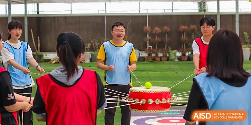 A male teacher or staff member at Suzhou Innovation Academy participating in a fun, active team-building game with a drum and small ball.