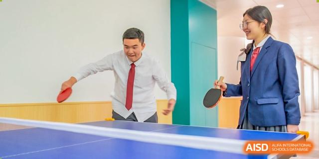 A male teacher or staff member from Suzhou Innovation Academy smiling while playing table tennis (ping pong) indoors.