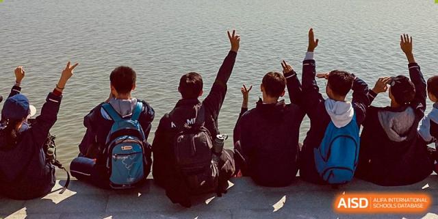 A group of Suzhou Innovation Academy students sitting on a ledge next to a body of water, raising their hands in a gesture of celebration or friendship.