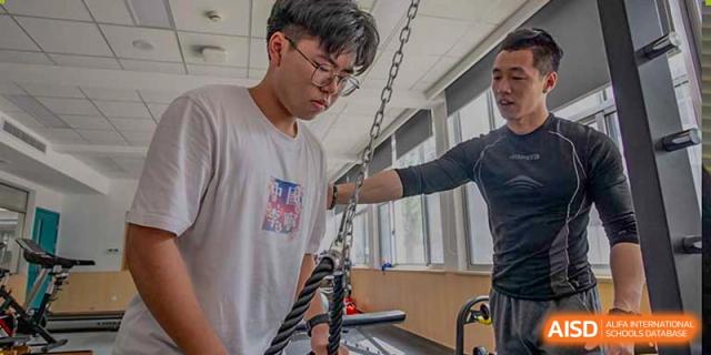 A male student from Suzhou Innovation Academy performing a strength training exercise using a cable machine or chains, assisted by a coach.