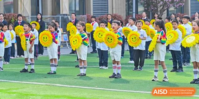 Young students at Walton International School Xuzhou standing on a green field in uniform, holding bright yellow sunflower props with smiley faces during an outdoor event or performance.