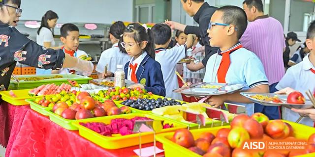 Students lining up to receive food, with a large, colorful display of fresh fruits in the foreground at the Walton International School Xuzhou cafeteria.