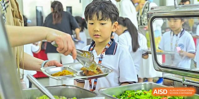 A young male student in uniform having his lunch tray served by a staff member in the cafeteria at Walton International School Xuzhou.