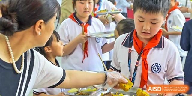 Students in uniform being served food by a gloved staff member from the buffet line at the Walton International School Xuzhou cafeteria.