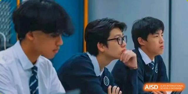 A male Wycombe Abbey School Hangzhou student with dark hair and glasses, wearing a navy sweater over a shirt and tie, looking away thoughtfully.