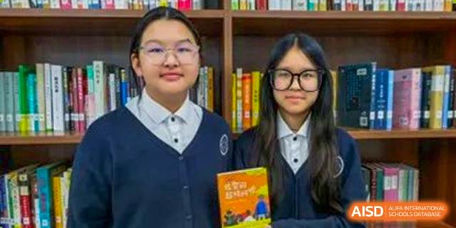 Two female Wycombe Abbey School Hangzhou students, wearing school uniforms and glasses, smiling while holding a book in a library.