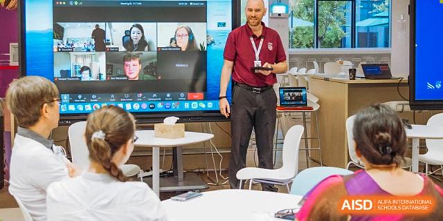 A male teacher at Xi'an Liangjiatan International School stands in a classroom giving a presentation with a digital screen behind him.