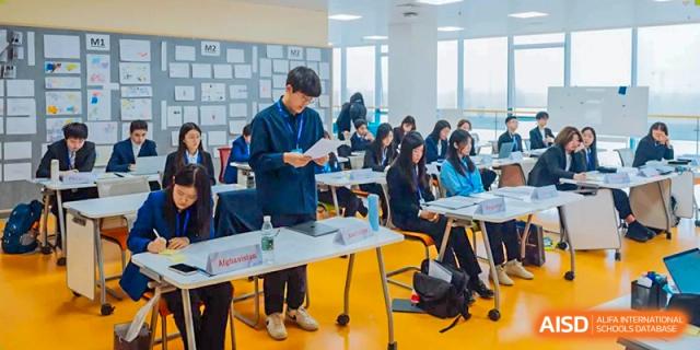 A student stands to present or address a group of other students sitting at tables in a bright classroom at Xi'an Liangjiatan International School.