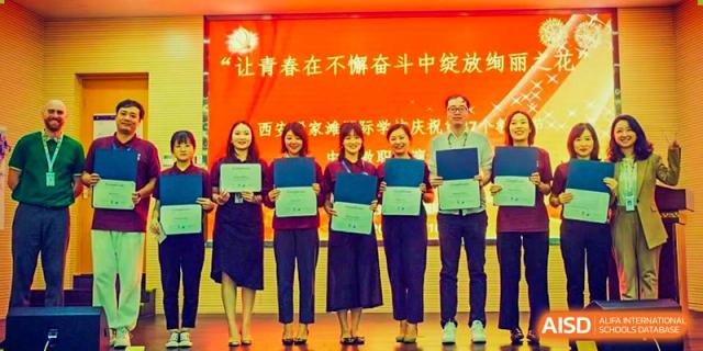 Students and teachers from Xi'an Liangjiatan International School pose on a stage, holding awards or certificates in front of a red banner with Chinese text.