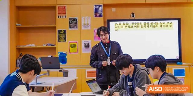 A teacher at Xi'an Liangjiatan International School assists a male student with his work on a laptop in a vibrant, poster-filled hallway or classroom.