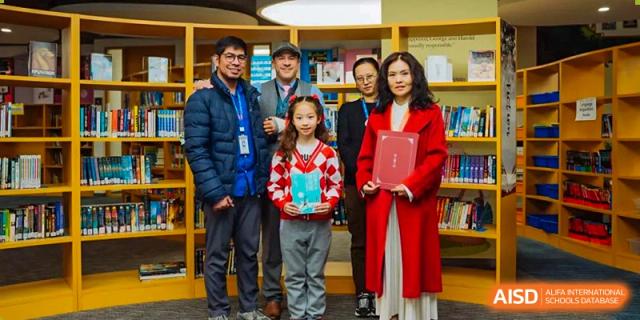 A group of students from Xi'an Liangjiatan International School stand with a female teacher, holding awards and standing in front of bookshelves.