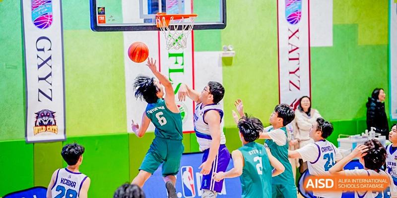 Close-up action shot of two players from Xi'an Liangjiatan International School teams, one in a green jersey and one in a purple and white jersey, reaching for a basketball near the hoop.