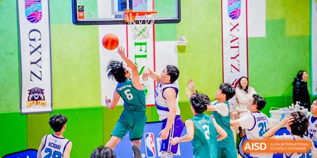 Close-up action shot of two players from Xi'an Liangjiatan International School teams, one in a green jersey and one in a purple and white jersey, reaching for a basketball near the hoop.