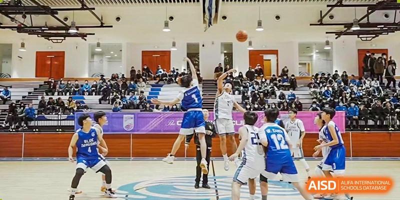 A player in a white and blue jersey for Xi'an Liangjiatan International School jumps high to shoot a basketball during a game in a packed gymnasium, with other players and spectators visible.